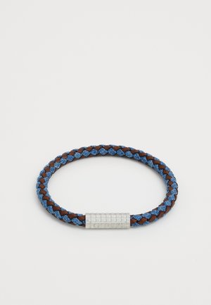 Geflochtenes Armband mit blauen und braunen Fasern, ausgestattet mit einem silbernen Metallverschluss, der den Markennamen GUESS trägt. Kreisförmig mit strukturiertem Oberflächenfinish.