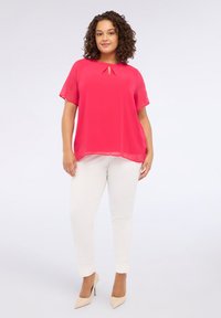 Kvinde med krøllet hår, iført en lys pink kortærmet bluse, hvide tætsiddende bukser og beige højhælede sko, der står foran en ensfarvet baggrund.