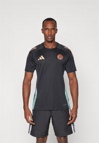 Sort træningsshirt med korte ærmer og turkise sidepaneler, orange logo og tre orange striber på skuldrene; parret med mørke shorts.