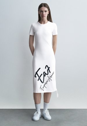 Naine seisab ühevärvilise seina taustal, kandes valget lühikeste varrukatega midi-kleiti, millel on "Ea7 Emporio Armani" logo, ning valgeid tossusid.