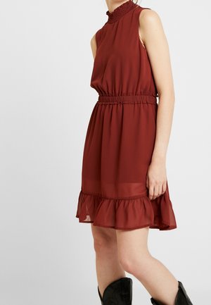 Robe rouge rouille sans manches avec col haut à volants, taille cintrée, ourlet à volants, portée avec des bottes noires mi-mollet sur fond clair.