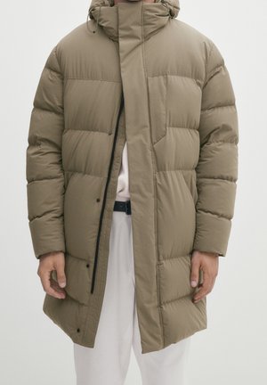 Mann trägt eine knieumspielende, olivgrüne Steppjacke mit Frontreißverschluss und Druckknöpfen über hellen Hosen vor einfarbigem Hintergrund.