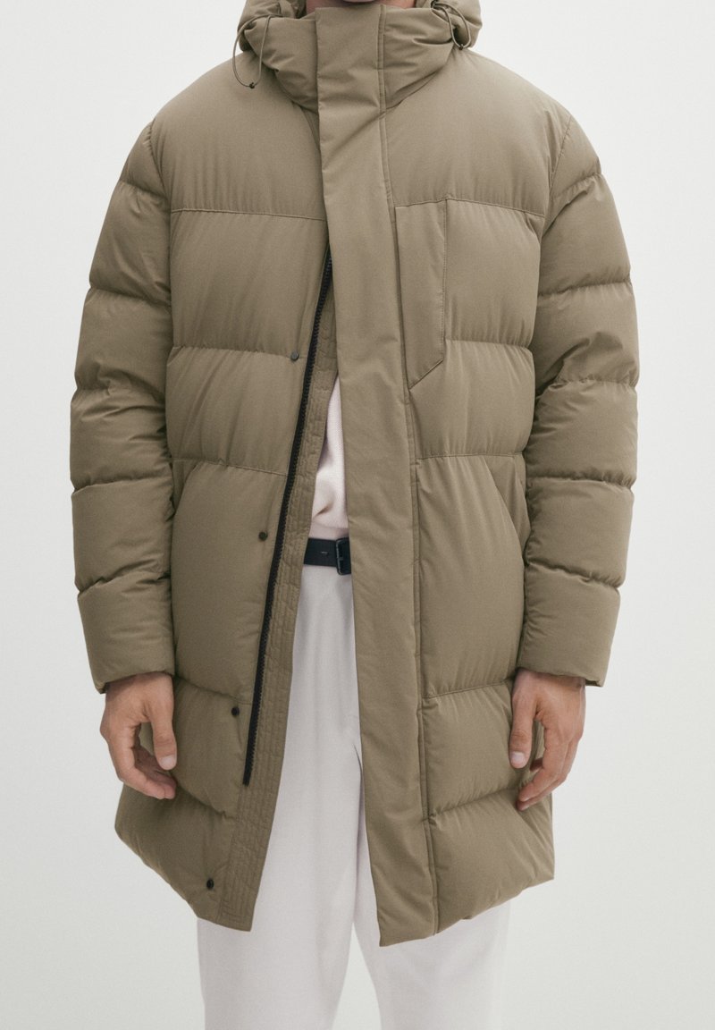 Homme portant une doudoune olive longueur genou avec fermeture éclair et boutons-pression à l'avant, sur un pantalon clair, sur un fond uni.