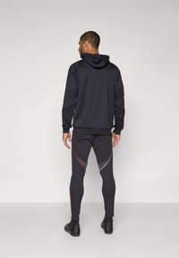 Conjunto de ropa activa para hombre en negro, con sudadera con capucha y pantalones ajustados con detalles en rojo y plata. Textura suave, diseño atlético.