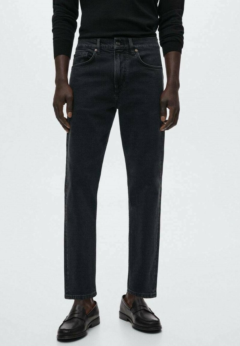 Homme portant un jean droit noir, un haut noir à manches longues et des mocassins en cuir noirs, debout devant un fond uni.