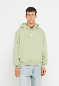 Felpa verde chiaro con tasca a marsupio, cappuccio con cordino e dettagli con logo. Tessuto morbido con una vestibilità comoda, abbinata a jeans in denim chiaro.
