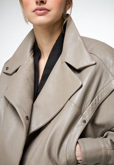 Femme portant une veste en cuir taupe surdimensionnée par-dessus un haut noir, main glissée dans la poche, lèvres fermées et cheveux attachés en queue de cheval lâche.