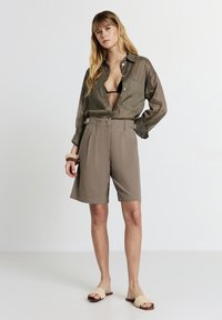 Donna in piedi con una mano sui fianchi, che indossa una camicia verde oliva con bottoni sopra una maglietta nera, pantaloni corti beige fino al ginocchio e sandali chiari senza lacci.