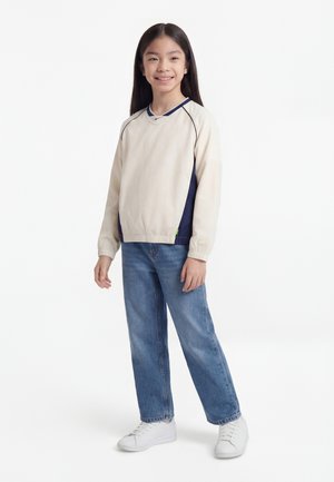 Lachend meisje met lang zwart haar staat en draagt een beige en marineblauwe trui, blauwe spijkerbroek en witte sneakers tegen een witte achtergrond.