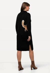 Donna che indossa un vestito a maglia nero fino al ginocchio con un design a coda di scoiattolo sulla schiena, abbinato a stivaletti con tacco in suede marrone.