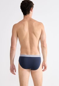 Slip blu navy con un elastico grigio chiaro in vita. Tessuto liscio, vestibilità aderente e copertura minima. La vista posteriore mostra dettagli di design semplici.