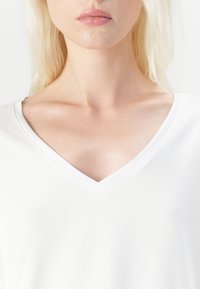 Bílá tričko s hlubokým véčkovým výstřihem. Měkká tkanina, lehká textura, minimální detaily šití kolem výstřihu. Důraz na jednoduchost.