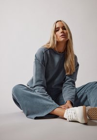 Avslappnad tvådelad outfit i mjukt blått tyg; består av en löst sittande långärmad topp och vida byxor, tillsammans med vita sneakers.