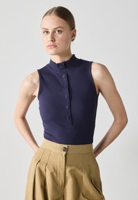 Top sin mangas de canalé en color marino con un alto cuello y detalle de botones en la parte delantera, combinado con pantalones de cintura alta de algodón color caqui con pliegues.