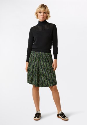 Schwarzes Langarmshirt mit strukturiertem Halsdetail, kombiniert mit einem knielangen grünen Rock mit geometrischem Muster, getragen mit schwarzen und beigen Sneakers.