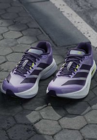Ζευγάρι μωβ και μαύρα παπούτσια για τρέξιμο Adidas Adizero τοποθετημένα σε εξαγωνική λιθόστρωτη επιφάνεια κοντά σε κράσπεδο δρόμου.