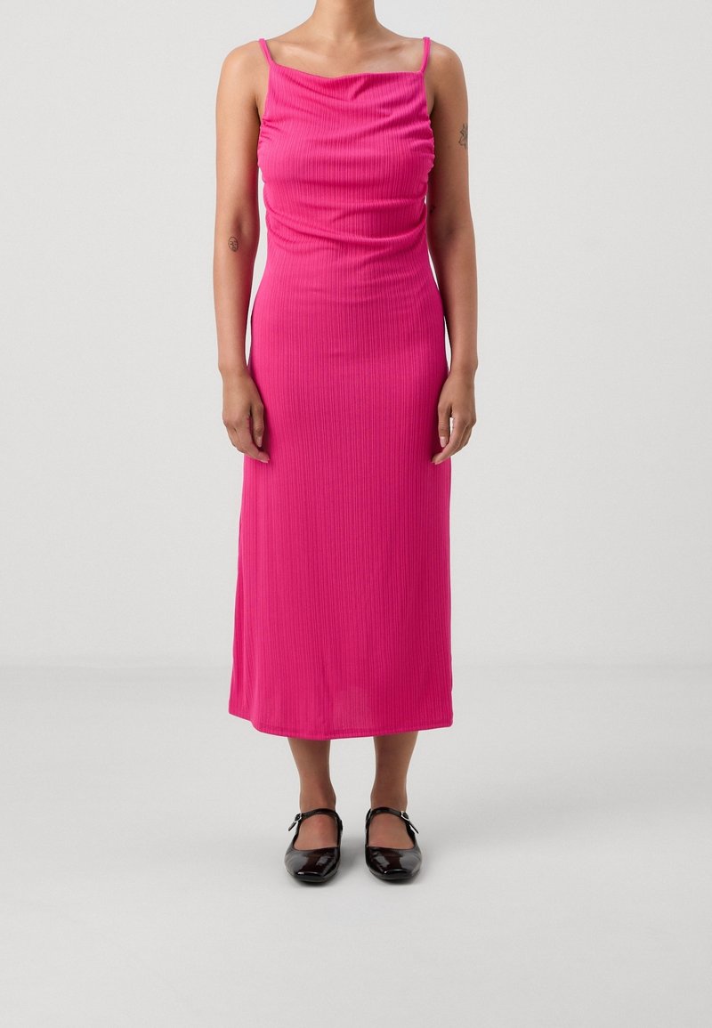 Femme portant une robe midi rose vif sans manches avec de fines bretelles et des chaussures Mary Jane noires, debout devant un fond blanc uni.