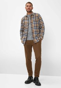 Bruine en blauwe geruite flanellen shirt over een grijze gebreide trui, gecombineerd met bruine broek en zwarte veterschoenen. Glad, gestructureerd materiaal.