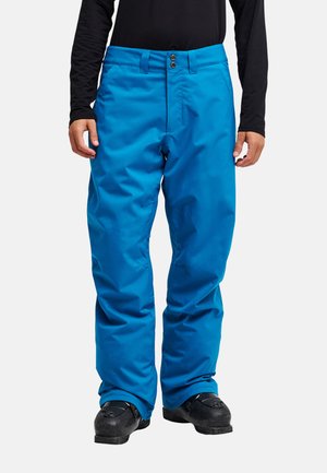 Person trägt leuchtend blaue Schneehose, schwarzen Langarmpullover und schwarze Schneestiefel und steht vor einem schlichten Hintergrund.