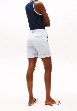 Personne portant un haut sans manches bleu marine, un short bleu clair à revers, et des sandales blanches à enfiler, debout les bras croisés, de dos.