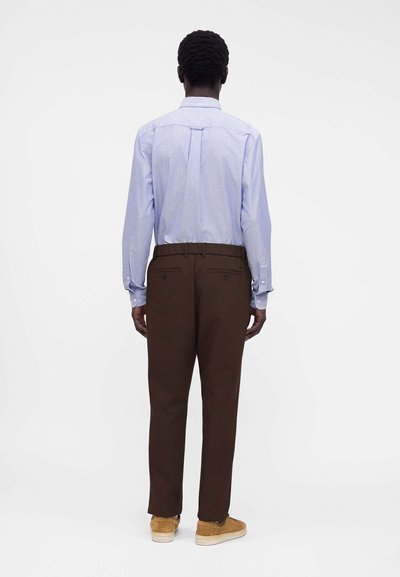 Homme de dos portant une chemise bleu clair à manches longues, un pantalon marron foncé et des baskets beige sur un fond blanc uni.