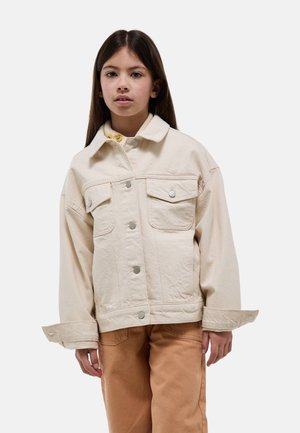 Fille aux longs cheveux bruns portant une veste beige clair boutonnée et un pantalon couleur sable, debout devant un fond blanc uni.