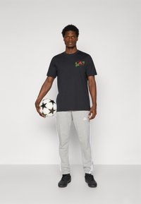 Sort t-shirt med rød og grøn design, grå sweatpants med hvide striber, holder en fodbold, sort fodtøj, ensfarvet baggrund.