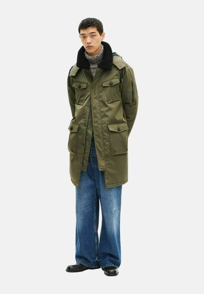 Oliwkowa parka z dużymi kieszeniami, zatrzaskami i teksturowanym czarnym kołnierzem, w połączeniu z luźnymi niebieskimi dżinsami i czarnymi butami.