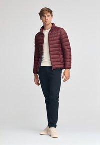 Burgundy puffjacka med horisontella sömmar, dragkedja och ståkrage, bärs över en krämfärgad tröja och matchas med mörka byxor och ljusa skor.