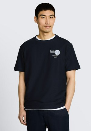 Mann trägt ein schwarzes T-Shirt mit kurzen Ärmeln, weißem Saum und kleinen Logodesigns auf der oberen linken Brust vor einfachem Hintergrund.