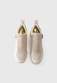 Une paire de baskets beiges à enfiler avec un dessus en tricot, des semelles blanches, des semelles intérieures dorées et des zips sur les côtés, vue d'en haut sur fond blanc.