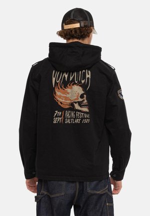 Personne portant une veste noire avec un crâne en flammes et le texte "Von Dutch Racing Festival Saltlake 1929" au dos, et un jean foncé.