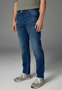 Slim-fit blauwe denim jeans met een middelhoge taille, vijf zakken en subtiele slijtage. Draagt met grijze sportschoenen.