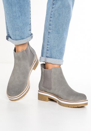 Personne portant un jean bleu clair retroussé et des bottines en daim gris avec des semelles épaisses en caoutchouc sur fond blanc.