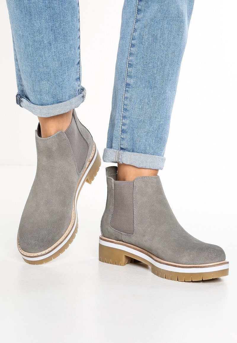 Personne portant un jean bleu clair retroussé et des bottines en daim gris avec des semelles épaisses en caoutchouc sur fond blanc.