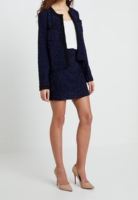 Veste texturée bleu marine avec bordures et poches noires, associée à une jupe ajustée assortie. Portée avec des escarpins beige clair.