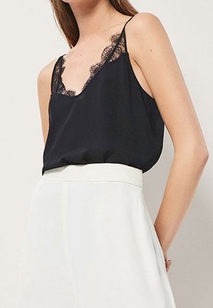 Woman wearing a black lace-trimmed camisole tucked into a high-waisted white skirt, standing with hands behind her back.