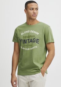Žalias medvilnės marškinėliai su apvaliu kaklu ir trumpomis rankovėmis, su spausdintu tekstu: "BLEND DENIM ORIGINAL VINTAGE EST. 1993." Lanksti tekstūra.