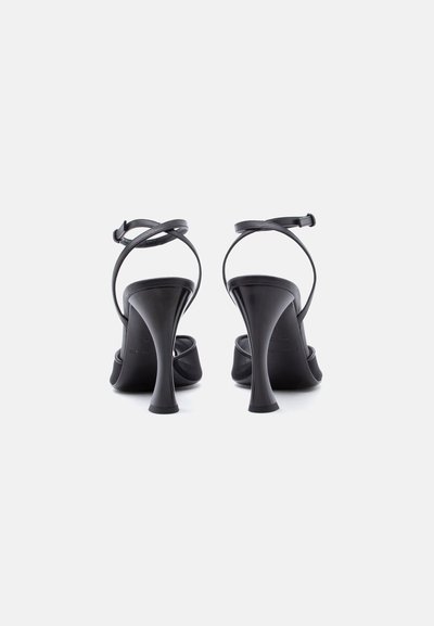 Paire de sandales à talons hauts noirs avec des brides fines autour de la cheville et des talons évasés, vues de dos sur un fond clair uni.