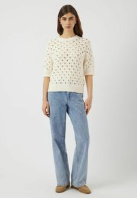 Krämfärgad plädtröja med öppen diamantsmönster, korta ärmar och ribbat fåll, tillsammans med ljusblå vida jeans och beige skor.