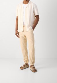 Lichtbeige shirt met korte mouwen en witte strepen, gecombineerd met lichtbeige tapered broek en bruine sandalen met dubbele banden.