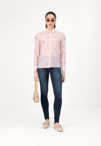 Light pink shirt with pink bow appliqués, button-up design, long sleeves, paired with dark jeans and beige flats, holding a small bag.