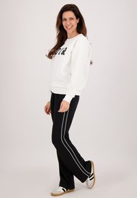 Witte sweatshirt met 'VIVA' in zwarte letters, gecombineerd met zwarte broek met witte zijstrepen en bruin-witte sneakers.