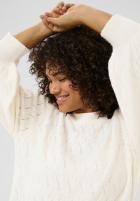 Pull en tricot blanc avec un motif en zigzag texturé, des manches larges et un col rond. La modèle a des cheveux bouclés avec les mains levées.