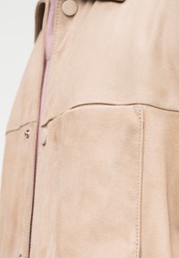 Μπεζ δερμάτινο μπουφάν από σουέντ με εμφανείς ραφές, κουμπιά σναπ και μπροστινή τσέπη, παρουσιάζοντας λεπτομερή υφή και υλικό σε κοντινό πλάνο.