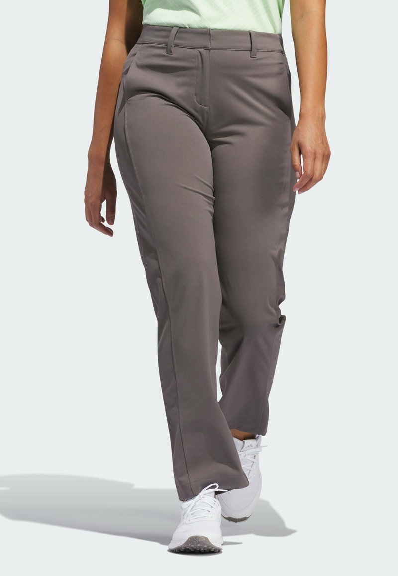 Personne marchant portant un pantalon gris droit, des baskets blanches et un haut vert clair sur un fond uni.