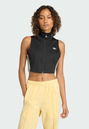 Donna con capelli ricci indossa un top corto nero senza maniche con zip e pantaloni da ginnastica gialli a vita alta con strisce laterali.