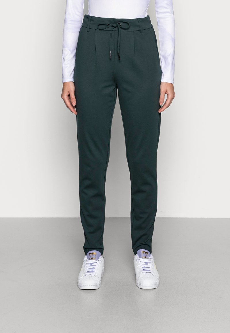 Femme portant un pantalon fuselé vert foncé avec taille à cordon, un haut blanc à manches longues et des baskets blanches, debout sur un sol gris.