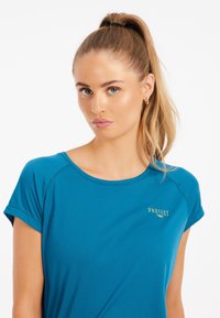Turkos färgad sport t-shirt med korta ärmar, rund hals och en guld "Protest" logotyp på bröstet. Slät textur, lätt design.