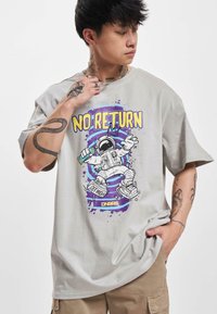 Lys grå grafisk t-shirt med et astronautdesign i lilla, gul og sort. Kortærmet og afslappet pasform med synlige tatoveringer.